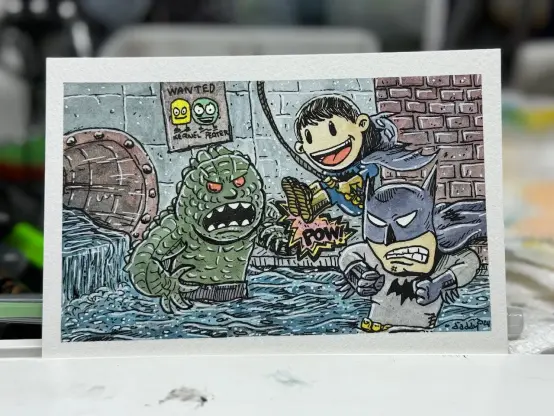 Cartoon-style drawing of Batman and young Batman fighting a large, green monster in a sewer. Young Batman is swinging mid-air, and Batman appears ready to punch. There's a "POW!" sound effect and a "WANTED" poster on the wall behind