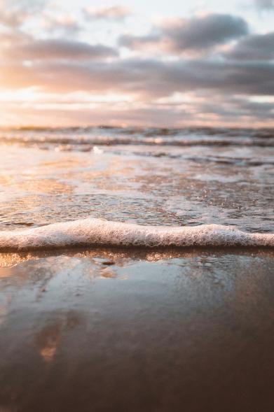 A close-up picture of the tide washing up on the beach, the sunrise, or sunset, just outside of the frame. There are clouds over the oceans and waves in the distance.