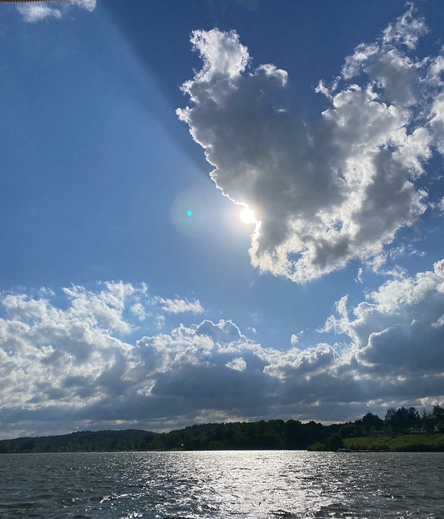 Color image of puffy cumulus clouds over a lake in Northeast Ohio, USA. Image shows the sun behind one cloud, that cloud then shadowing other clouds, leaving the sky in differing shades of blue and dark blue by the shadowing, the shorline a mile off, and the water glistening.