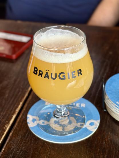 A glass of beer labeled "BRÄUGIER" on a wooden table, placed on a blue coaster.