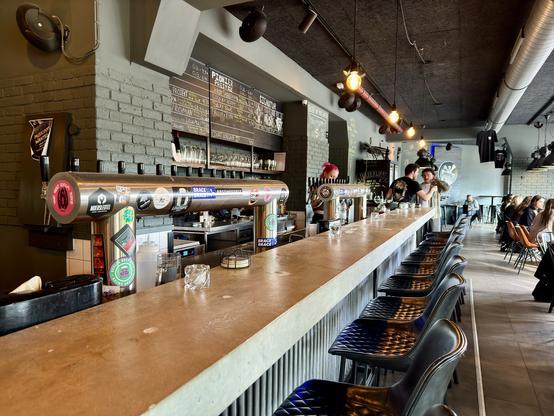 A modern bar with high stools lined along a long counter. Multiple beer taps are visible along with a menu on the wall. Bartenders are serving customers, and patrons are seated at tables in the background. The ambiance is casual with contemporary decor.