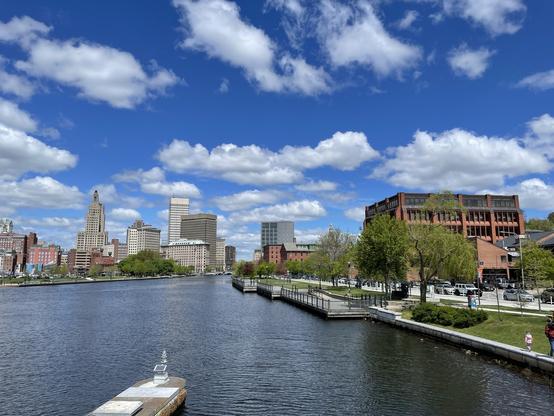 The Providence city skyline across a river with a dock on the right and a walkway along the waterfront. Clear blue sky with scattered clouds, good vista of buildings including Superman.