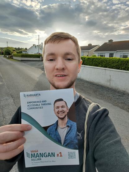 A selfie of me holding my leaflet with the sun in the evening sun and houses in the background