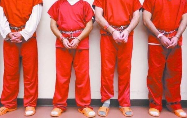 Cropped heads out - 4 men, handcuffed with chains around their waists, standing against a white wall, wearing orange prison uniforms, with white socks & orange sandals.