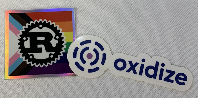 A Rust sticker with a Pride Flag in the background next to a sticker with the oxidize logo