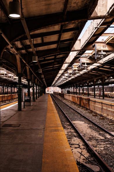 A color photograph showing a long rail platform and tracks heading away from the camera. An arched overhead covering protects the platform with opening over the tracks to allow light in, and the parallel tracks on either side have the same. In the distance, past the end of the platform, buildings are visible in the bright midday sun. Yellow, rubberized warning strips line the platform edge. A black iron fence separates the adjacent track. A parked train is visible four tracks to the right on the edge of the frame.