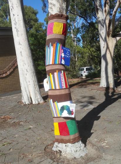 Tree wrapped in crochet with shelves of crocheted books in lots of colors. One shelf shows the cover of Cat in the Hat full and it is open. Another shows the Very Hungry Caterpillar.