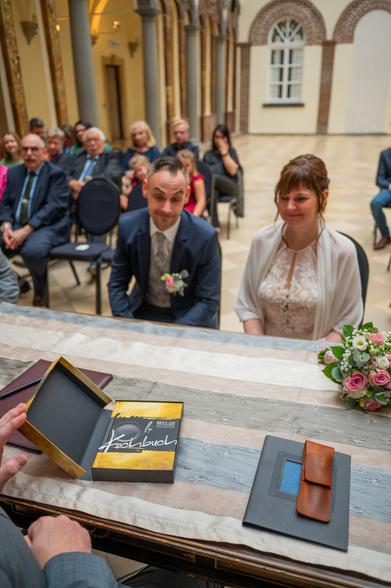 Das Foto zeigt eine Trauung auf einem Standesamt, der Beamte überreicht den beiden ein kleines Präsent der Stadt und zwar ein hübsch eingepacktes Kochbuch. 
Daneben ist noch das Stammbuch zu sehen, vor dem Tisch sitzt das Brautpaar, dahinter die Gäste.