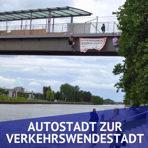 Eine Brücke über einem Wasserweg. Es haben sich 2 Menschen abgeseilt und zwischen sich ein Banner gespannt auf dem steht "Achtung umbauarbeiten, Transformation, Konversation und Vergesellschaftung. Autostadt zur Verkehrswendestadt.