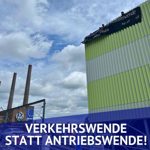Verkehrswende statt Antriebswende und verkehrswendestadt.de steht auf einem Banner, dass von einem grünen Gebäude hängt. Dahinter zwei Menschen. Links im Bild eine Fabrik mit Schornsteinen und VW-Logo