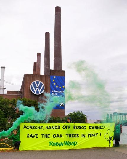 Menschen die ein Banner halten auf dem steht "porsche hands off Bosco D'arneo Save the oak trees in italy. dahiner eine Fabrik mit Schornsteinen und VW-Logo. Es ist grüner Rauch zu sehen.