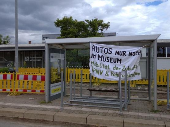 Eine Bushaltestelle vor dem Automuseum. Daran hängt ein Banner "autos nur noch im Museum. Mobilität der zukunft ÖPNV & Fahrradstraßen
