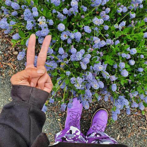 Flashing hand peace sign with my left hand, looking down at my purple camo pants & purple boots, standing in front of large patch of blueish purple soap bush flowers.