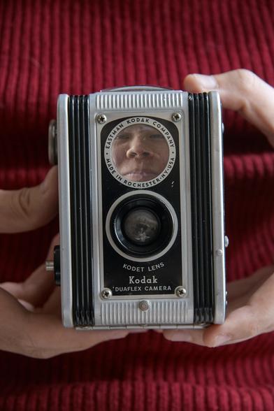 A closeup portrait of a Kodak Duaflex Camera, as a portrait. A person’s face appears reflected in the reflex lens, looking almost like there is a tiny person trapped inside. The camera is held in front of their red sweater, highlighting the contrast between the silver and black camera body and the background.