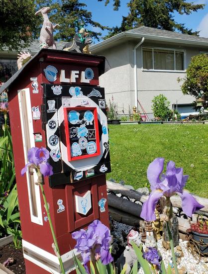 My little free library in front yard. Purple irises in front of it. My library box has environmental & social justice stickers plastered all over it. There's dinosaur, Baby Yoda, Teenage Mutant Ninja Turtle figurines on the roof of it.