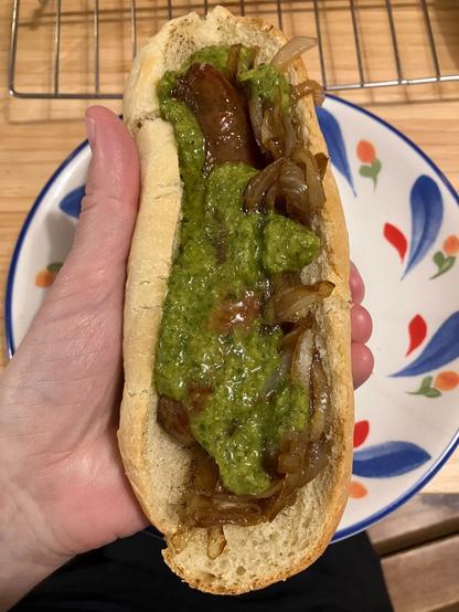 Choripán: A sausage on half of a grilled mini baguette with caramelized onions and green chimichurri sauce held in a hand over a plate