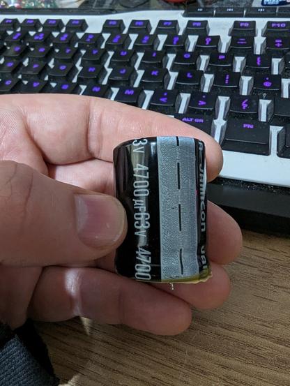A photo of a large capacitor being held. The text on it reads 4700uf 63v. There is a small amount of dried yellow residue around the base of the capacitor
