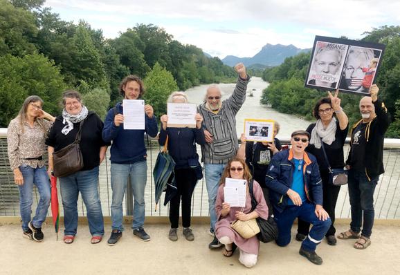 Un groupe de dix manifestants, composé de six femmes et quatre hommes, posent sur la passerelle du pont Frédéric Mistral. La moitié brandit des affiches ou documents en faveur de la libération de Julian Assange. En arrière-plan coule la Drôme entre ses berges verdoyantes qui laissent apparaître, au loin, les Trois Becs de la Forêt de Saoû.