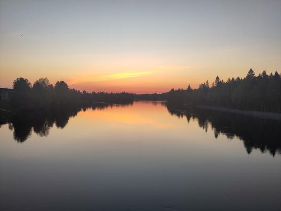 A flat river in pink-orange glow from a sun just under the horizon. There's trees on the banks, and some hints of houses.