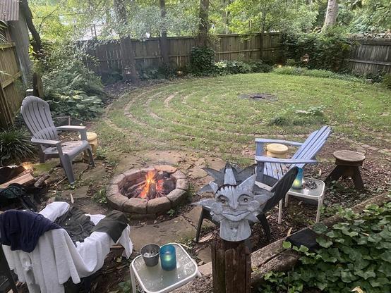 A back yard with a seven-circuit labyrinth in the background. In the lower left, a fire pit with a fire, surrounded by Adirondack chairs and small tables. It is a scene of calm and seclusion.