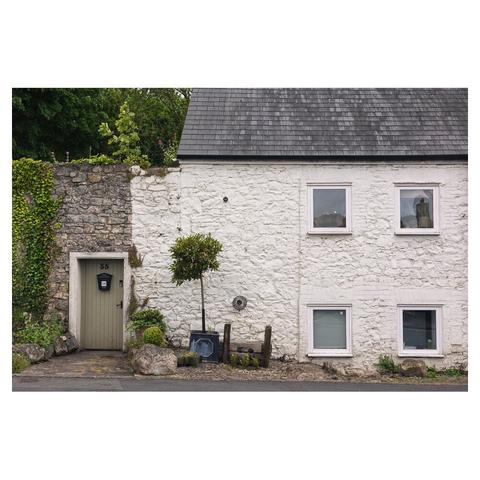 a front on picture of a white, stone walled house with a grey slate roof and 4 equally placed windows. on the left is a plain stone wall with a sage green door. there are stocks outside the front door