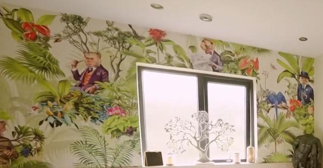 A wall  photographed at an angle. There is a window in the middle. There is very unusual wallpaper. It has tropical flowers and ferns with cameos of creatures in old fashioned clothes.