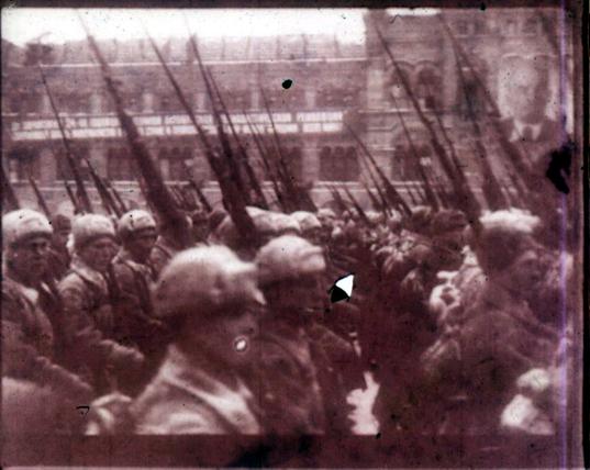 A group of Soviet soldiers march in a parade. A large portrait of Lenin is visible on the building in the background.