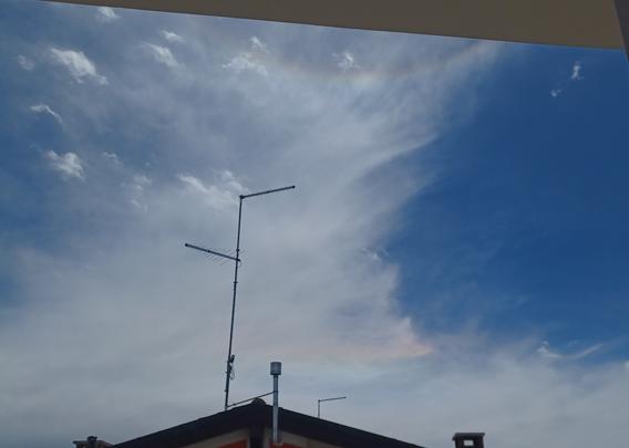 Blue sky with white cirrus clouds above the roof of a house. Iridescent colors can be seen in the clouds just above the roof. Iridescence also forms an arc similar to an upside-down rainbow in the upper part of the image.