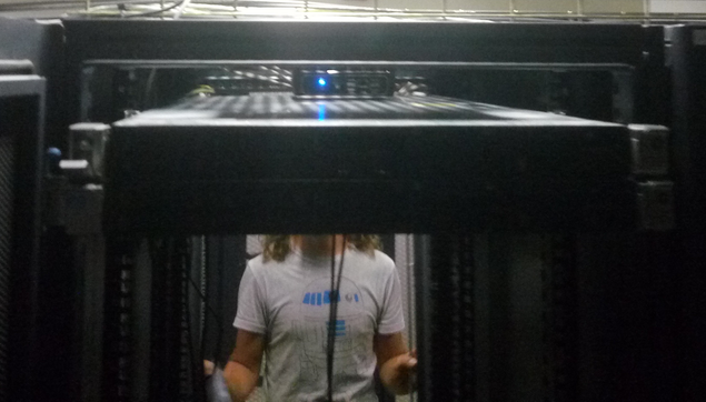 Dans un datacenter, on voit en avant plan un serveur dans une armoire et en arrière plan un humain dont le torse apparait dans l'encadrement de l'armoire. La tête de l'humain est masquée par le serveur.