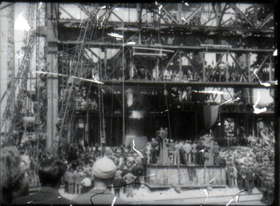 A black and white still that shows a large number of people in and around a building covered in scaffolding.