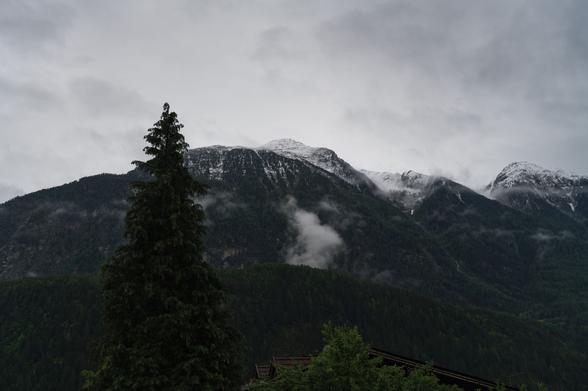 Foto einer in den Gipfelbereichen verschneiten Bergkette, der Himmel bewölkt und grau, kleinere Wolken auch im Bereich der Bewaldung.