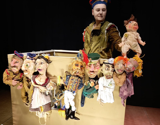Die Darstellerin mit 9 Puppen, eine hält sie in der Hand, die anderen hat sie an ihre Box gehängt. Sie trägt ein Soldatenoutfit.