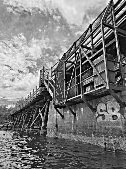 Black & white photo.
Selkirk Trestle Bridge. Shot from waters below.
Graffiti that says SB is on cement foundation. An old power station platform is attached to foundation with fenced cage around it. A locked off fenced gate & stairwell lead down to platform. You can see bridge upper profile & wooden trestles below.