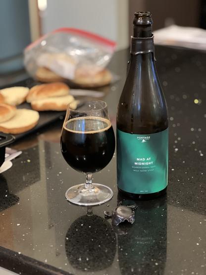 A 750ml dark brown bottle with a green label has a tulip glass of beer to its left. in the background are signs of prep for dinner.