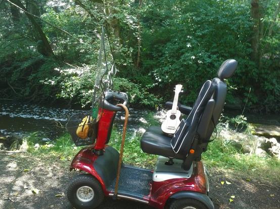 My red mobility scooter with front windshield & my yellow purse in front black metal basket, with my yew wood cane hanging from handlebar & my ukulele & ukulele bag on seat. It is parked beside the river.