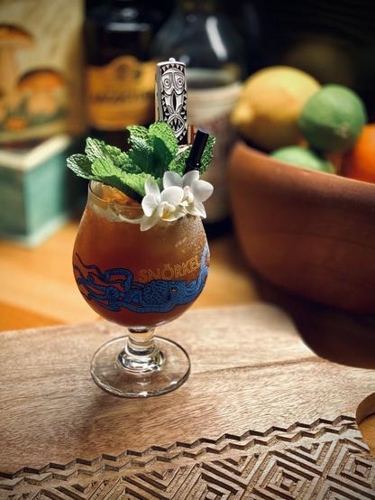 brown cocktail with white orchid mint sprig and white False Idol swizzle stick garnish and surfside sips black bamboo glass straw in a Jester King snörkel glass sitting on an etched wooden board with lemon lime and orange citrus in a wooden bowl, skippers finest demerara rum, house of angostura amaro di angostura and porcini mushrooms depicted on a box in the background
