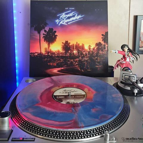 A Seastorm variant vinyl record sits on a turntable. Behind the turntable, a vinyl album outer sleeve is displayed. The front cover shows a road surrounded by palm trees leading to a city in the background around sunset.