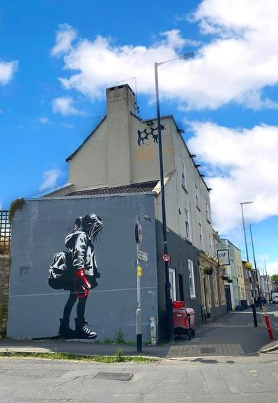 Streetartwall. The mural of a little girl was sprayed onto a gray exterior wall using stencils. The girl is mainly depicted in shades of gray.  She has a ponytail hairstyle, wears an anorak, tights, sports shoes and a school rucksack on her back.
She walks with her head down and has a red boxing glove on her hand. She seems to be on her way to school. Title: "Stop bullying" (The photo shows the corner house, a pub and the roads and traffic signs. Above the mural you can see a painted cow on the wall of the house. It is the trademark of the pub behind it).