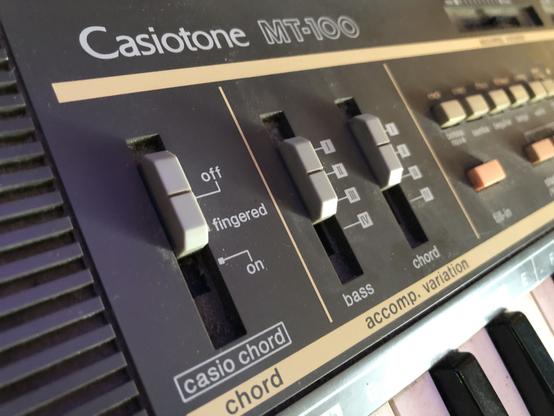A long shot down a vintage synthesizer made by Casio, the front panel reads: "Casiotone MT-100" above several sliders and buttons.