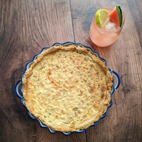 A quiche in a pie dish next to a cocktail on a dark wooden background. Little pieces of sweetcorn, watermelon rind and jalapeño are visible in the quiche. The dish is fluted, with two wee curved handles and a painted-blue top. The cocktail is a pink drink in a low-ball glass, filled with ice, with a lime wheel and watermelon slice garnish.