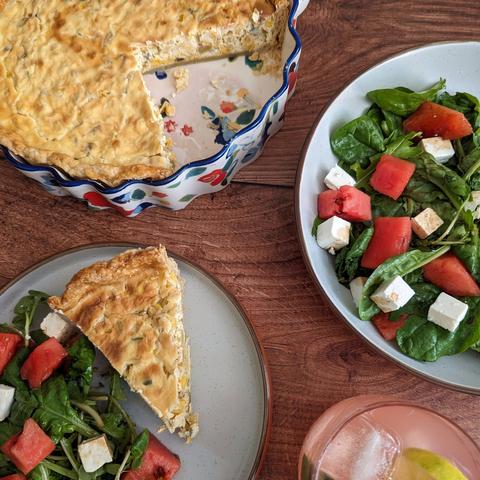Food and drink on a dark wooden background. In the top right is the quiche from the first photo, with a couple of slices cut out. To the right is a green-leaf salad with cubes of watermelon and feta and balsamic vinegar. To the bottom right is the cocktail. To the bottom left is a plate with a slice of the quiche and some of the salad.