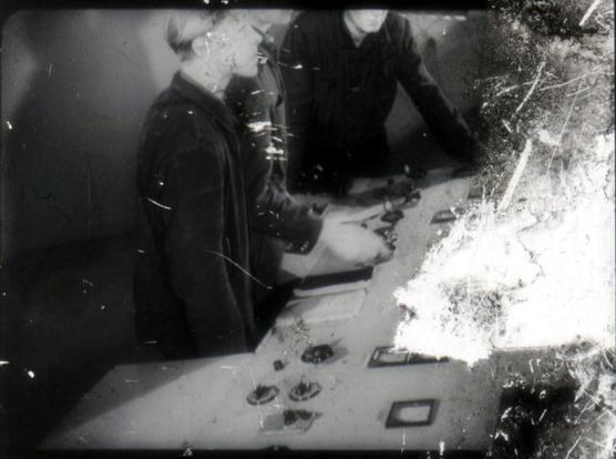 A black and white still of two people working at a control desk with switches and dials.