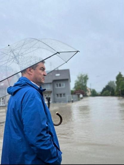Markus Söder steht mit einem Regenschirm auf einer überfluteten Straße im Hochwassergebiet