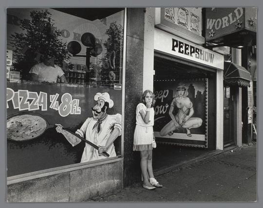 This image depicts a scene from Ed van der Elsken's series "Meisje buiten een peep- en pornoshow op het Damrak," captured between 1983 and 1990. The scene takes place on the street in front of a store that appears to sell pizza, as indicated by the signage in the background. In the foreground, there is a person standing with their back facing the camera, looking out onto the street. Behind this individual, there are two advertisements. One features an illustration of a chef slicing a pizza, while the other shows a woman wearing a short dress and high heels, suggesting a provocative or adult nature to the content displayed in the store's window. The photograph has an aged appearance due to its vintage quality, which is common for images from that period.