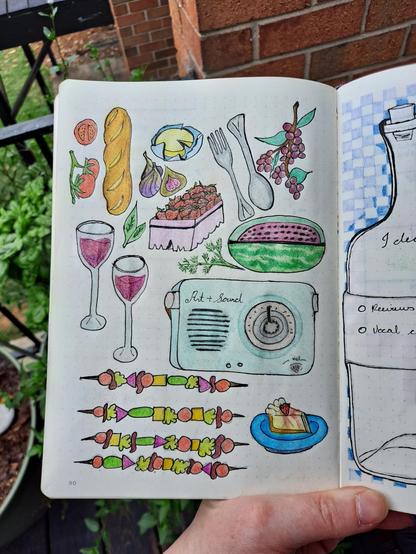 The image shows my bullet journal held by me. The page is filled with colorful, hand-drawn illustrations of various items which may be found at a picnic or on an outing to an outdoor market. These items include:

- A baguette
- A slice of watermelon
- A small container of strawberries
- A couple of tomatoes on the vine
- A few figs
- A paper wrapper of creamy yellow cheese
- A couple of wine glasses with red wine
- A vintage radio labeled "Art + Sound"
- A set of utensils (fork and spoon)
- A sprig of grapes
- Several skewers (kebabs) with colorful pieces of vegetables and meat
- A slice of cheesecake with a strawberry and drizzled in strawberry glaze on a blue plate

My bullet journal is held outdoors near some potted plants and a brick wall, on the deck of my apartment. The potted plants here serve as my garden. The illustrations are neatly arranged and are part of the creative aspect of my journaling experience. As an artist, I often will add some creative features to my journal, be it artworks of this nature, stickers, or photographs.