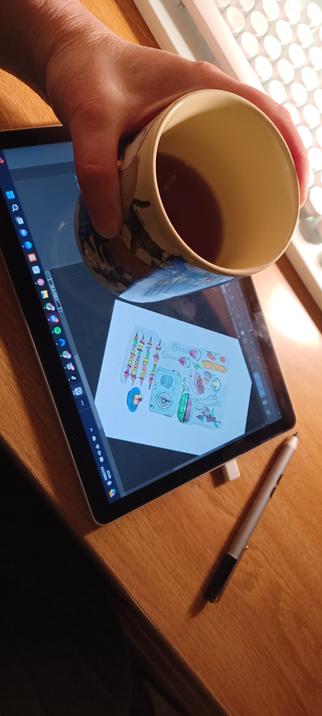 The image shows my hand holding a mug of blueberry crumb cake flavored tea above a tablet on a wooden desk. The tablet screen displays a digital art project with colorful drawings of various items found at a picnic, such as a cordless radio with a vintage looking appearance, food, and drinks, arranged in a hexagonal layout. The tablet is running Clip Studio Paint, a graphics and illustration software, for creating digital art and design work. I am working on digitizing an image I drew in pencil and ink. Once it's finished, I will upload it to my Redbubble shop as a sticker design. A stylus pen for the tablet, is lying on the desk to the right of the tablet. In the background, there is a portion of a white and mint colored keyboard. The image was taken indoors, in a low-light setting, late at night. 

To learn more about the image I'm working on, check out the alt text in the other photo in this same post. Thank you for taking an interest in my work.