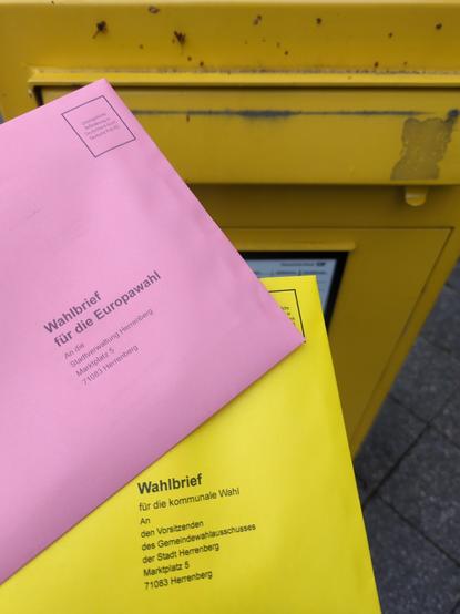 Zwei Briefumschläge, einer rosa, einer Gelb mit der Aufschrift "Wahlbrief für die Europawahl" bzw. "Wahlbrief für die kommunale Wahl". Im Hintergrund ein gelber Briefkasten.