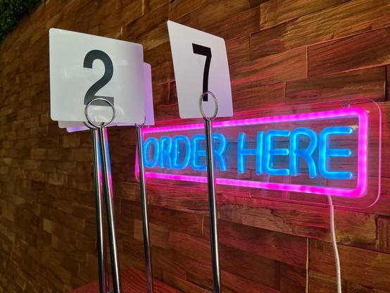 On the left of the photo there are some plastic table signs on a chrome stand and on the right in the background is a neon sign in blue and pink that says Order Here.
