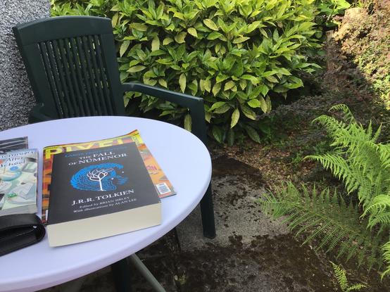 A white garden table and green garden chair beside a grey harled wall and on the other sides masses of large green leafy plants. On the table are a number of magazines and books, including a large print The Fall of Numenor by Tolkien.