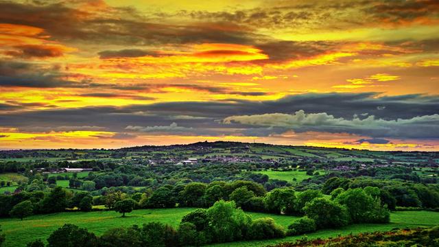 A vibrant sunset over a lush, green countryside with rolling hills and scattered buildings. The sky is filled with dynamic clouds, painted in shades of yellow, orange, and purple.